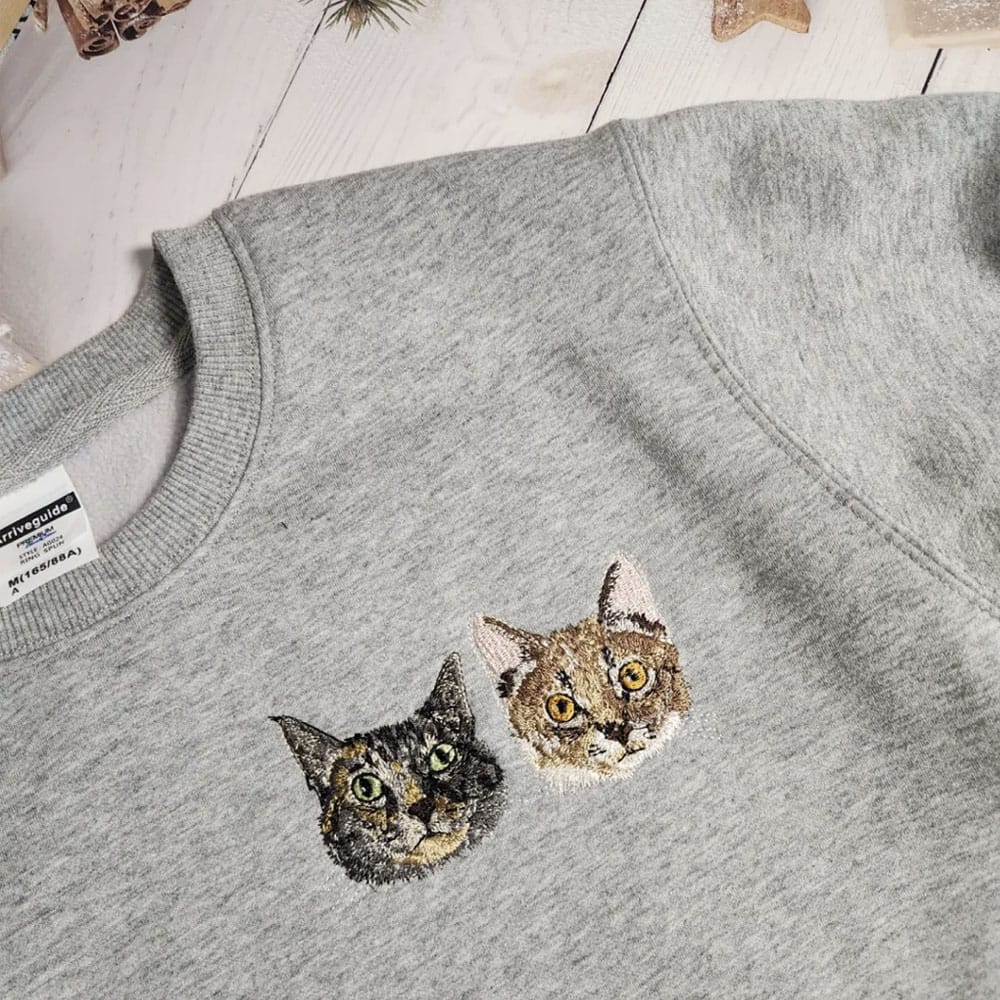 Custom embroidered pet portrait featuring two detailed cat heads stitched on a gray crewneck sweatshirt.