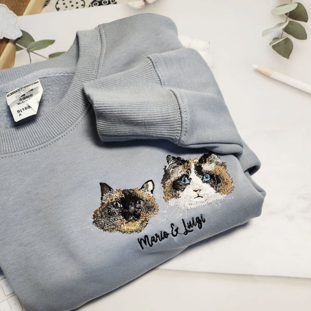 A folded light blue crewneck sweatshirt featuring custom embroidered portraits of two cats—a Siamese and a Ragdoll—with the personalized names "Mario & Luigi" stitched below