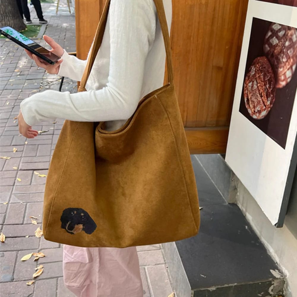 Person holding a brown suede bag with a embroidered dog face on a sidewalk.