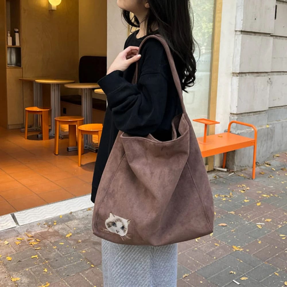 Person holding a suede bag with a embroidered cat face on a sidewalk.