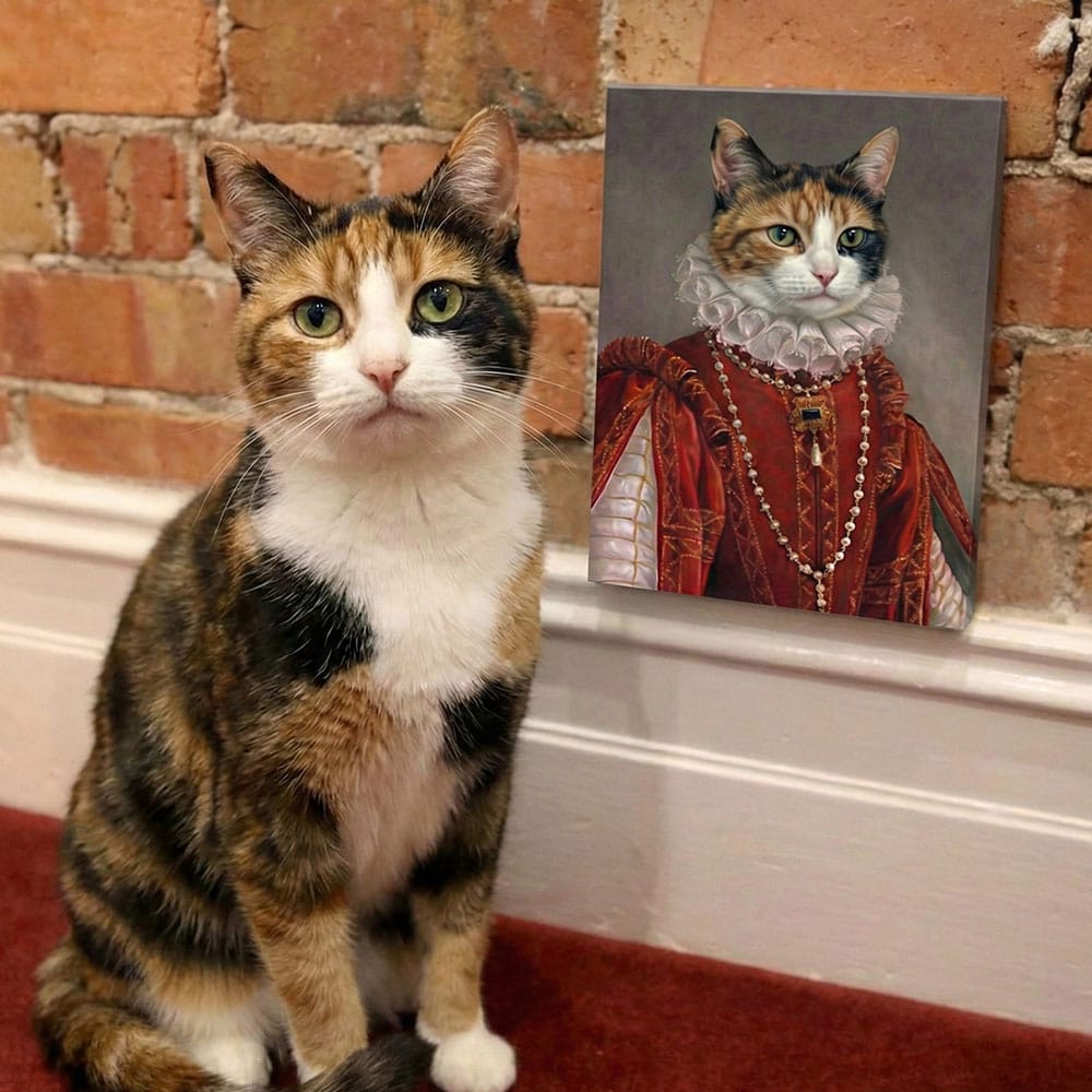 Calico cat sitting next to a painting of a cat in historical attire against a brick wall.