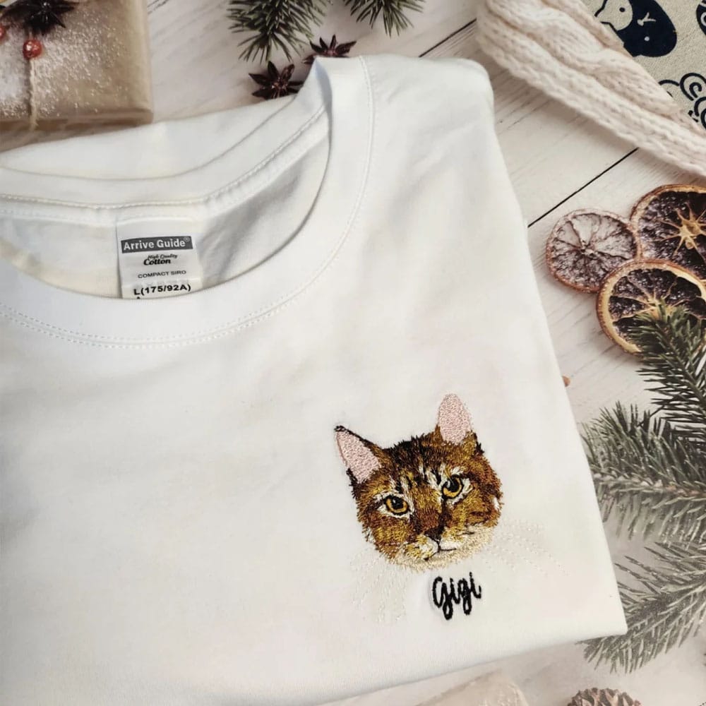 White tshirt with a embroidered cat face and cat name on it.