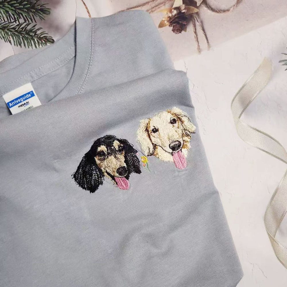 A light blue short-sleeve T-shirt laid flat on a white surface, featuring two embroidered dog faces on the chest.