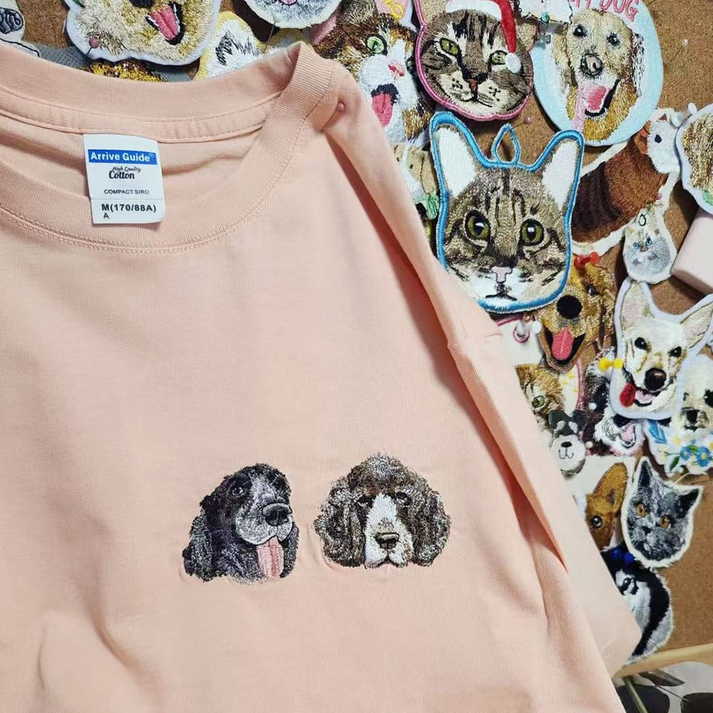 A light peach-pink crewneck sweatshirt featuring embroidered dog faces on the front, displayed on a cork board surrounded by dog and cat stickers.