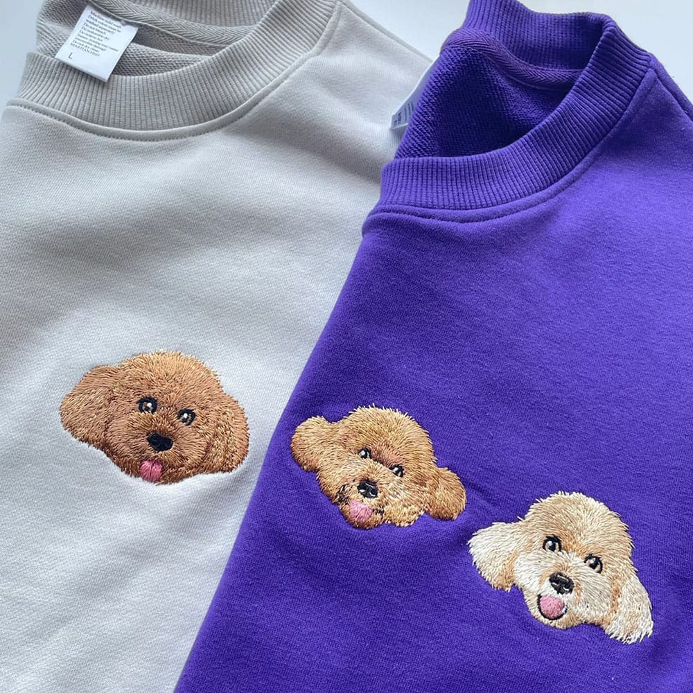 Two sweaters, one white and one purple, with embroidered dog faces on a light gray background.