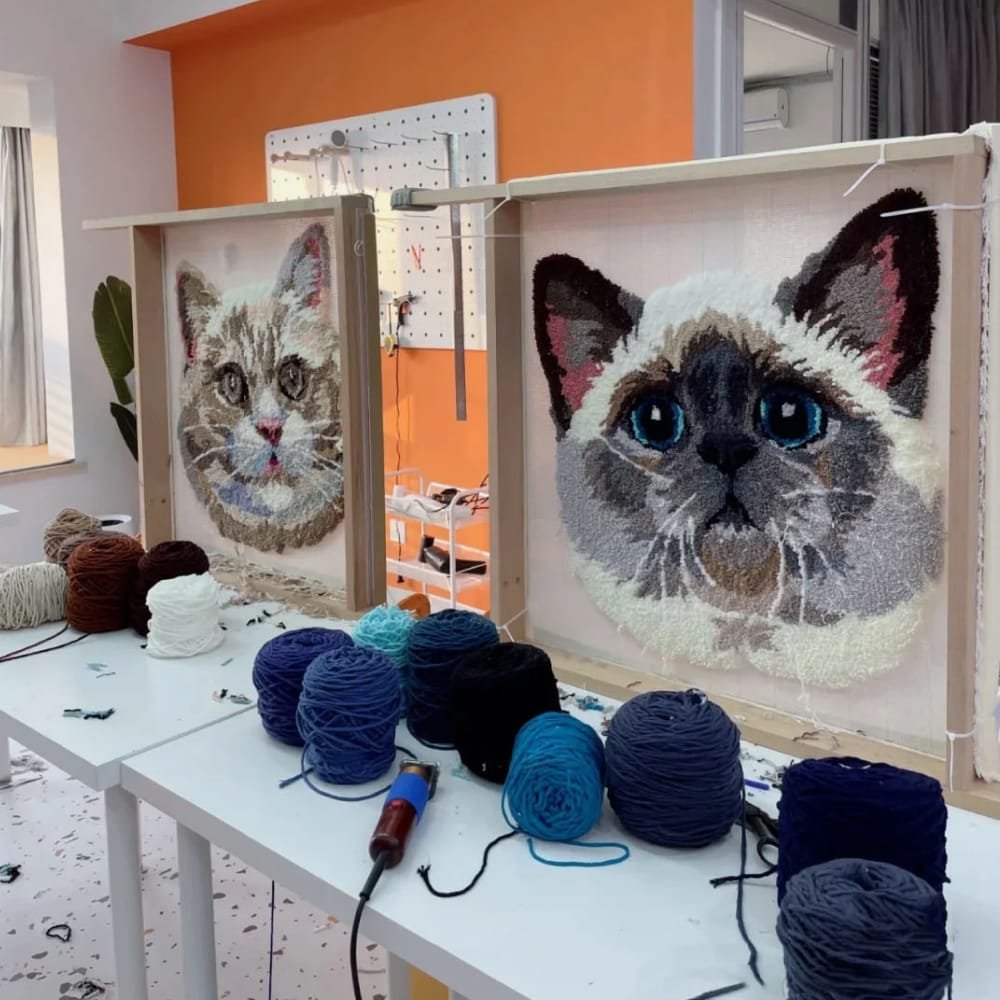 Two framed needlepoint cat portraits on a table with yarn and tools.