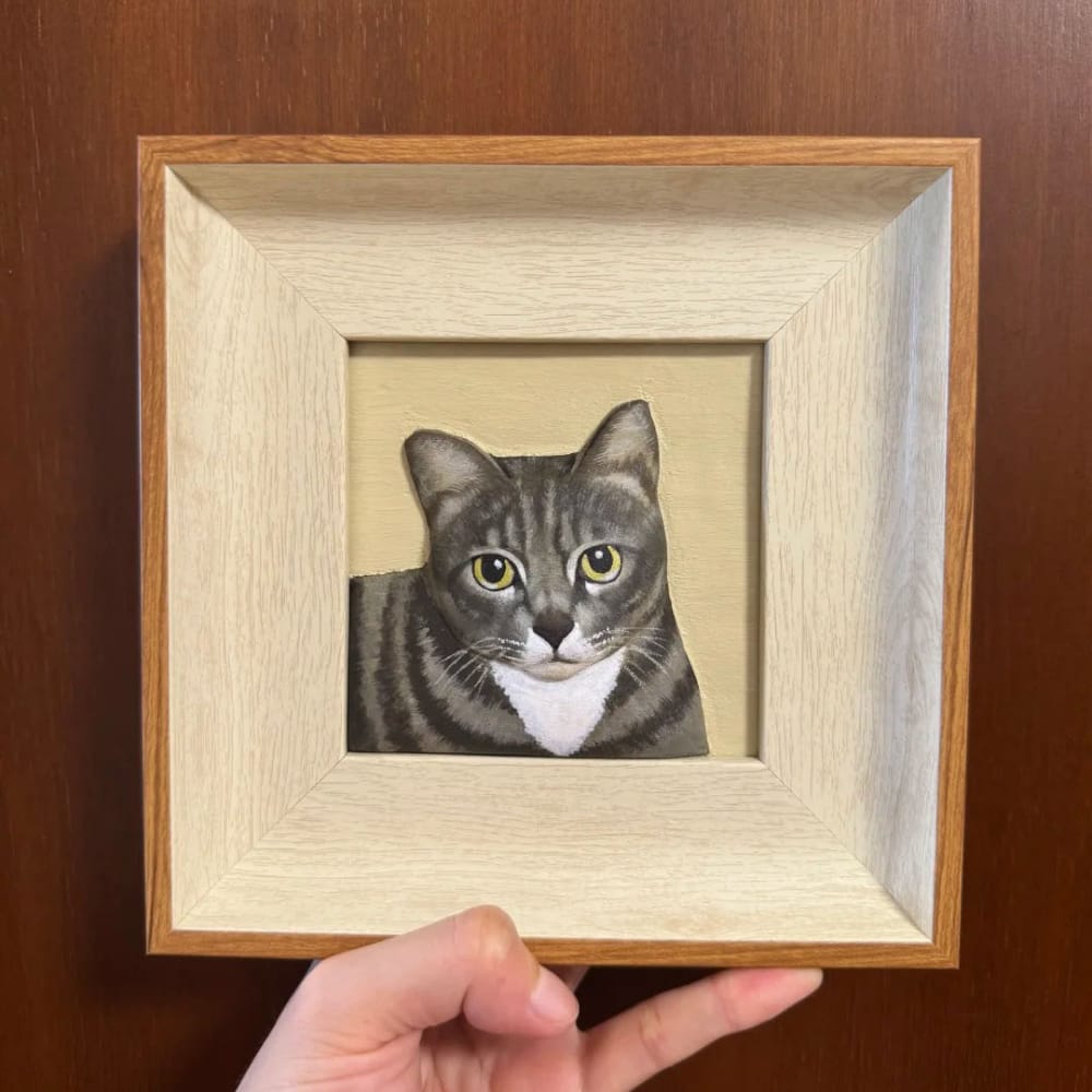 A custom pet portrait wood carving of a grey tabby cat, held inside a light wood frame.