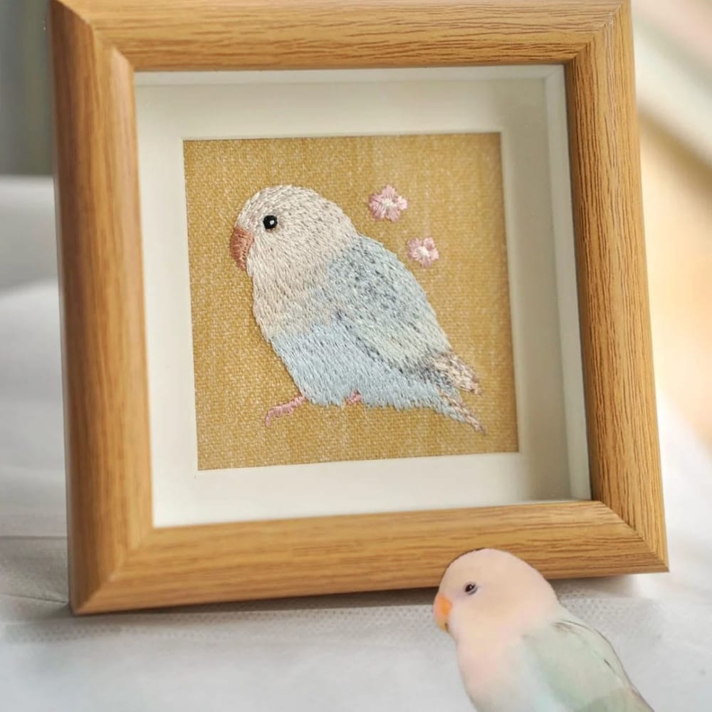 Framed custom embroidered bird portrait of a light blue lovebird in a light wood frame.