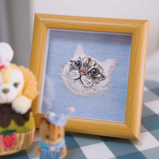 A framed custom embroidered portrait of a fluffy cat's face on a light blue background, set in a light wood frame.