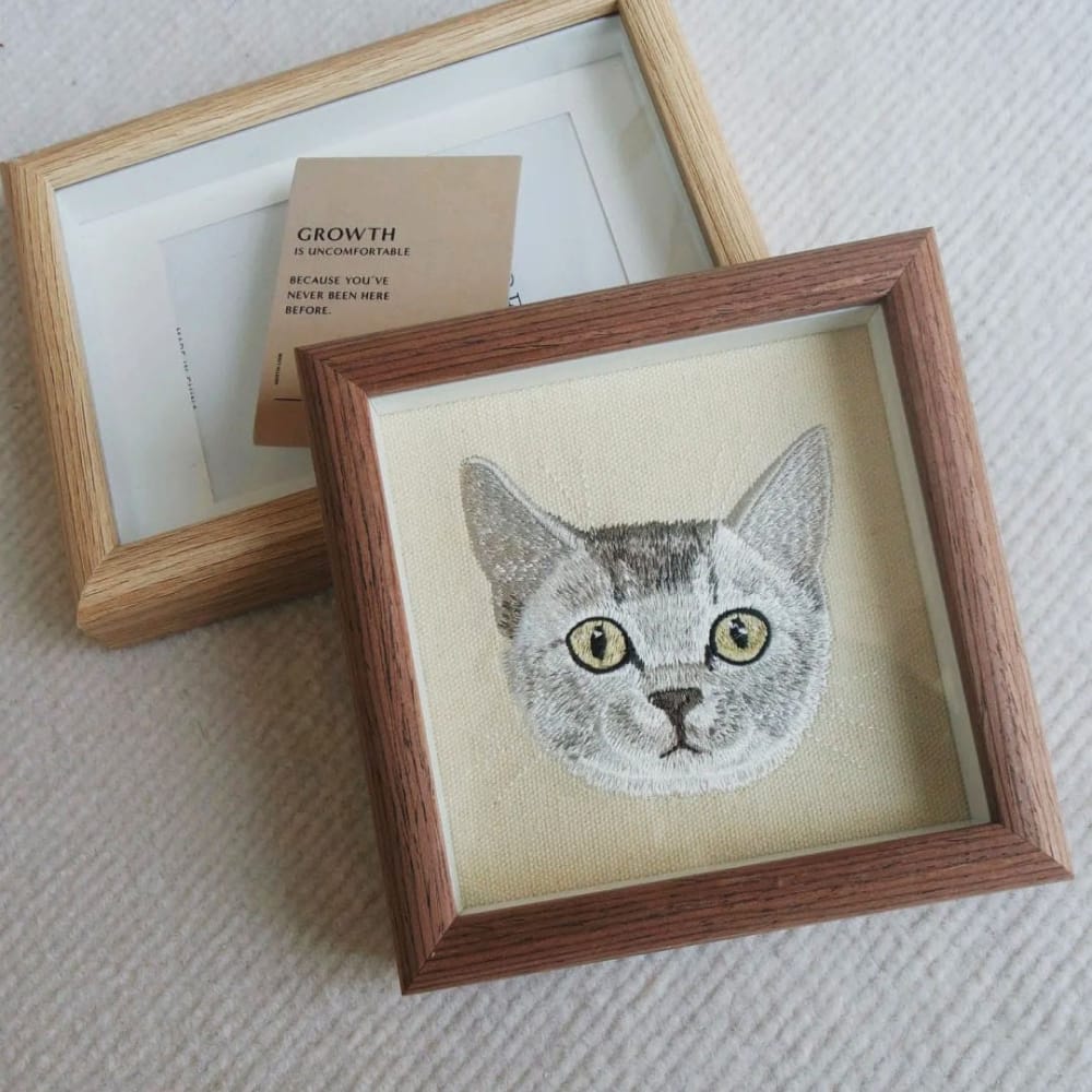 A framed custom embroidered cat portrait, showing a grey cat's face in a wood frame.