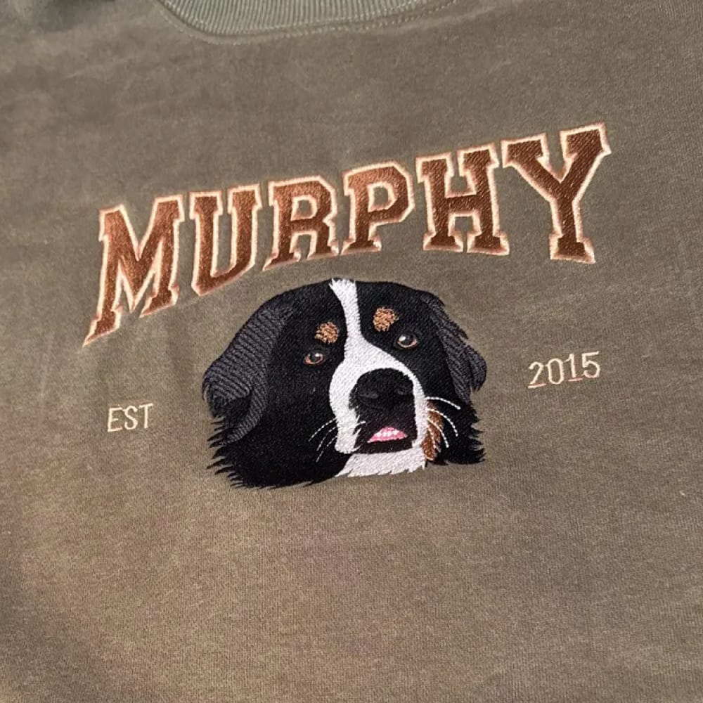 A brown sweatshirt featuring a custom embroidered dog portrait with personalized text in a varsity style font.