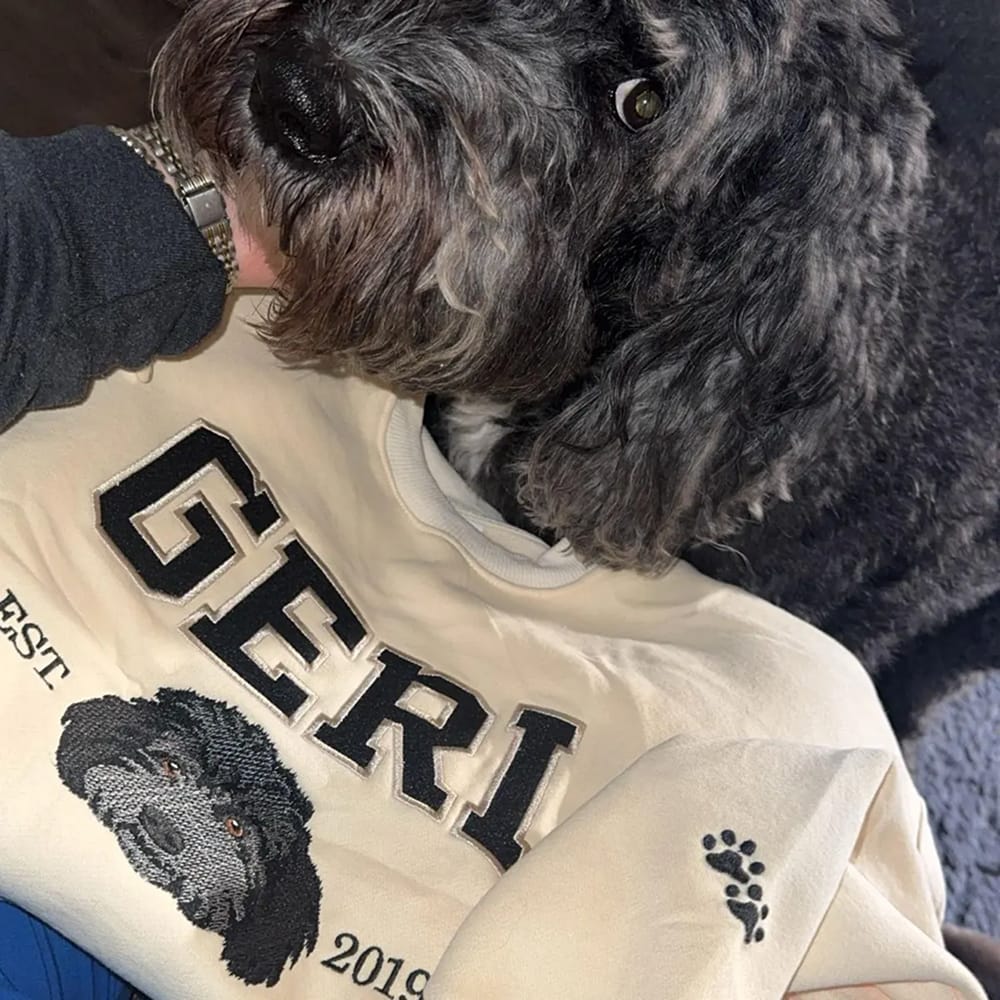 A cream sweatshirt featuring a custom embroidered dog portrait with personalized text in a varsity style font.