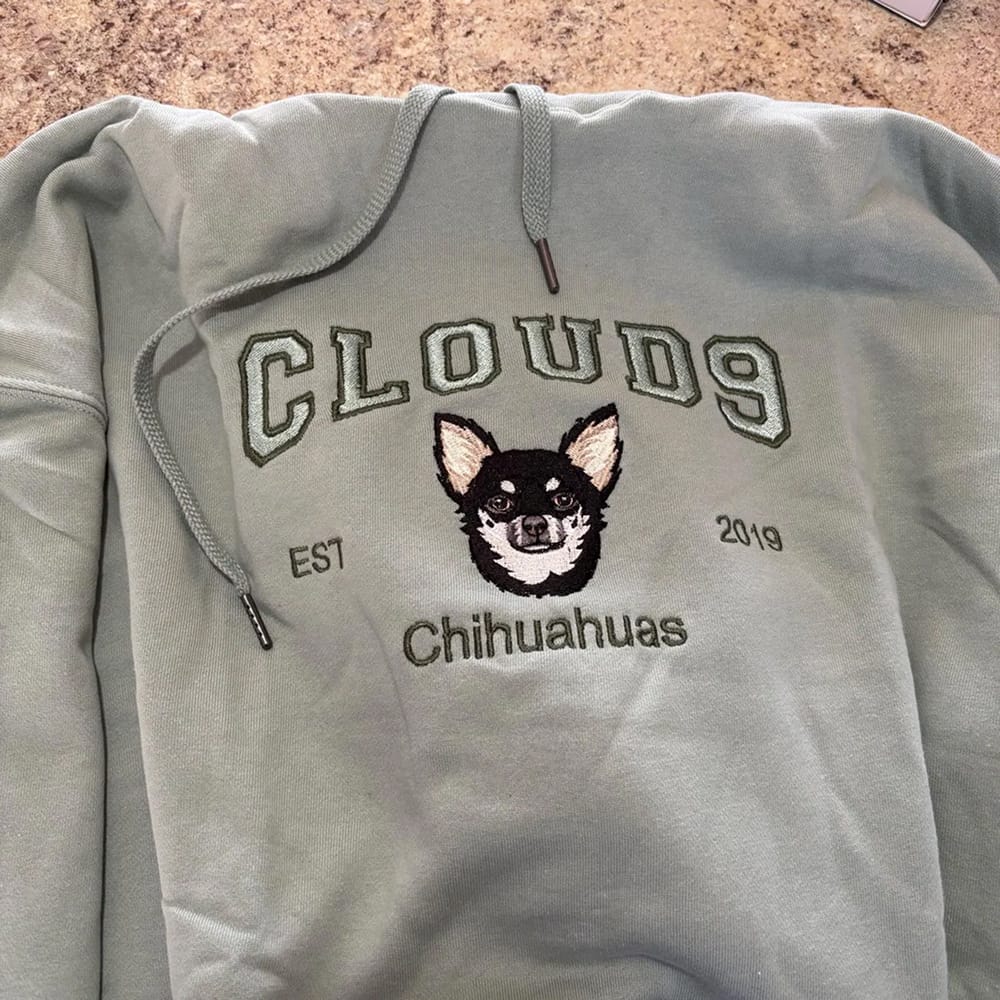 A gray sweatshirt featuring a custom embroidered dog portrait with personalized text in a varsity style font.