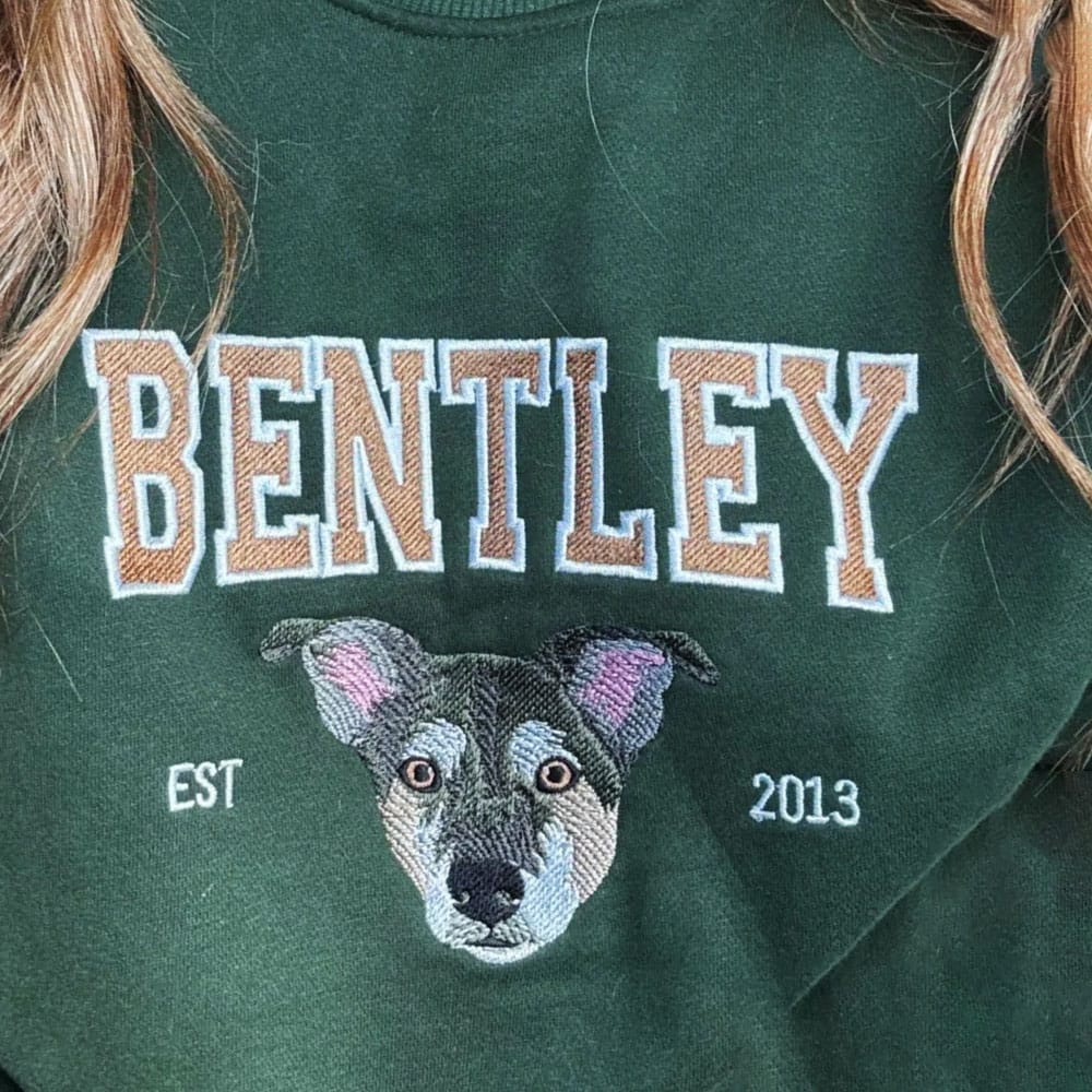 A green sweatshirt featuring a custom embroidered dog portrait with personalized text in a varsity style font.