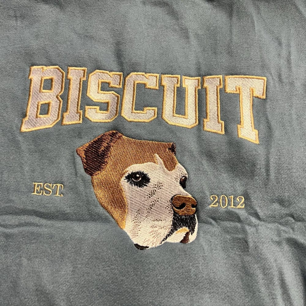 A sweatshirt featuring a custom embroidered dog portrait with personalized text in a varsity style font.